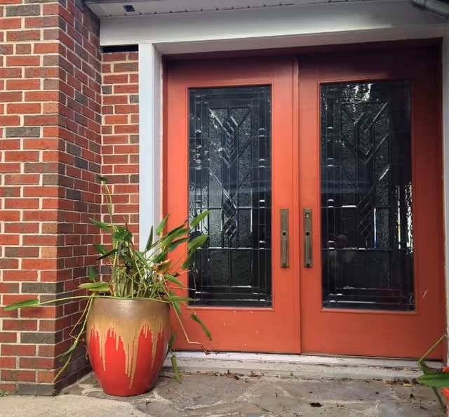 Double red decorative glass front doors set in a brick exterior with a potted plant beside them.