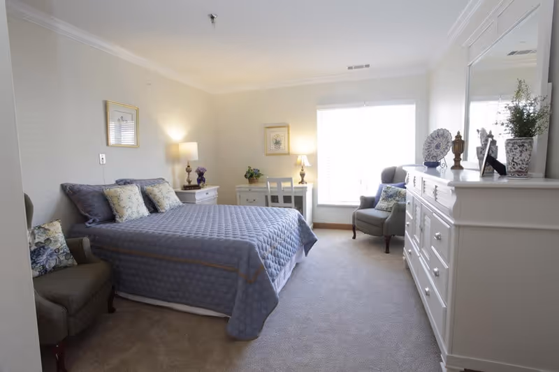 Bright furnished bedroom with a bed, two armchairs, dresser and a window letting in natural light.