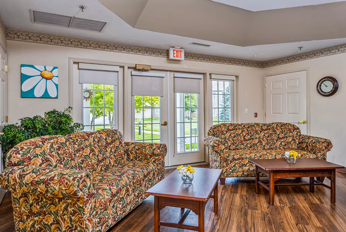 A bright sitting room with two floral-patterned sofas, wooden coffee tables with small flower vases, and glass doors leading outside.