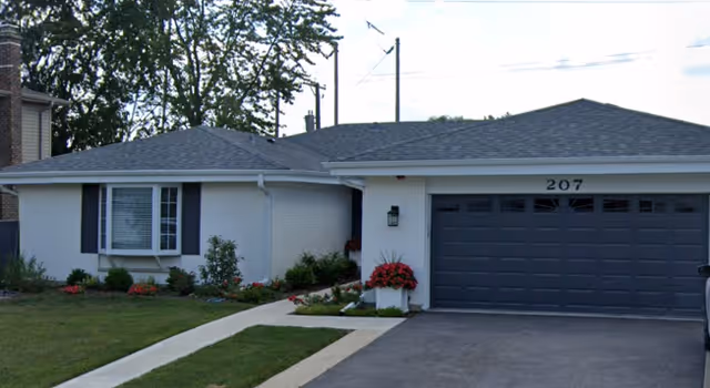 Single-story residential building with a gray roof, white exterior walls, and black shutters. There is a driveway leading to a double garage with the number 207 above it. The front yard has a well-maintained lawn, a concrete walkway, and flower beds with red flowers and shrubs.
