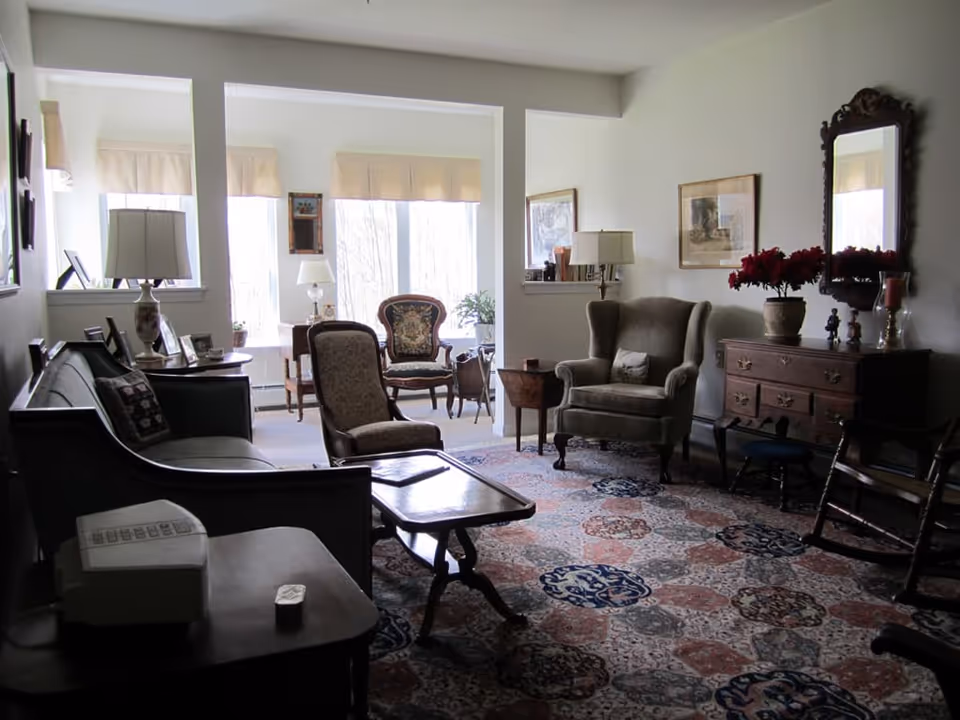 A cozy living room with vintage furniture including a sofa, armchairs, a wooden coffee table, a side table with a lamp, a wooden dresser with a mirror, and decorative items such as framed pictures and a vase with red flowers. The room has large windows with beige valances letting in natural light and a patterned area rug covering the floor.