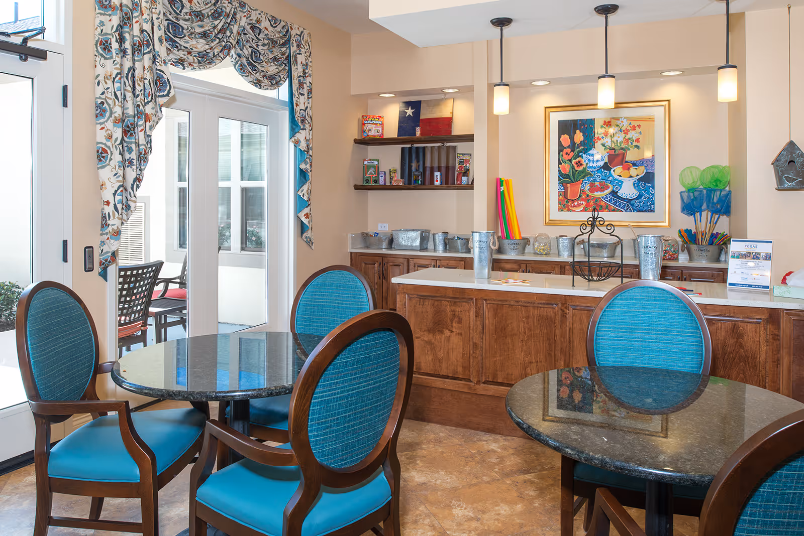Interior view of a common area with round tables and blue cushioned chairs. There is a counter with wooden cabinets and various decorative items, including a colorful painting of flowers and fruit on the wall. Shelves hold games and other items, and there are pendant lights hanging from the ceiling. A glass door with floral curtains leads to an outdoor seating area.