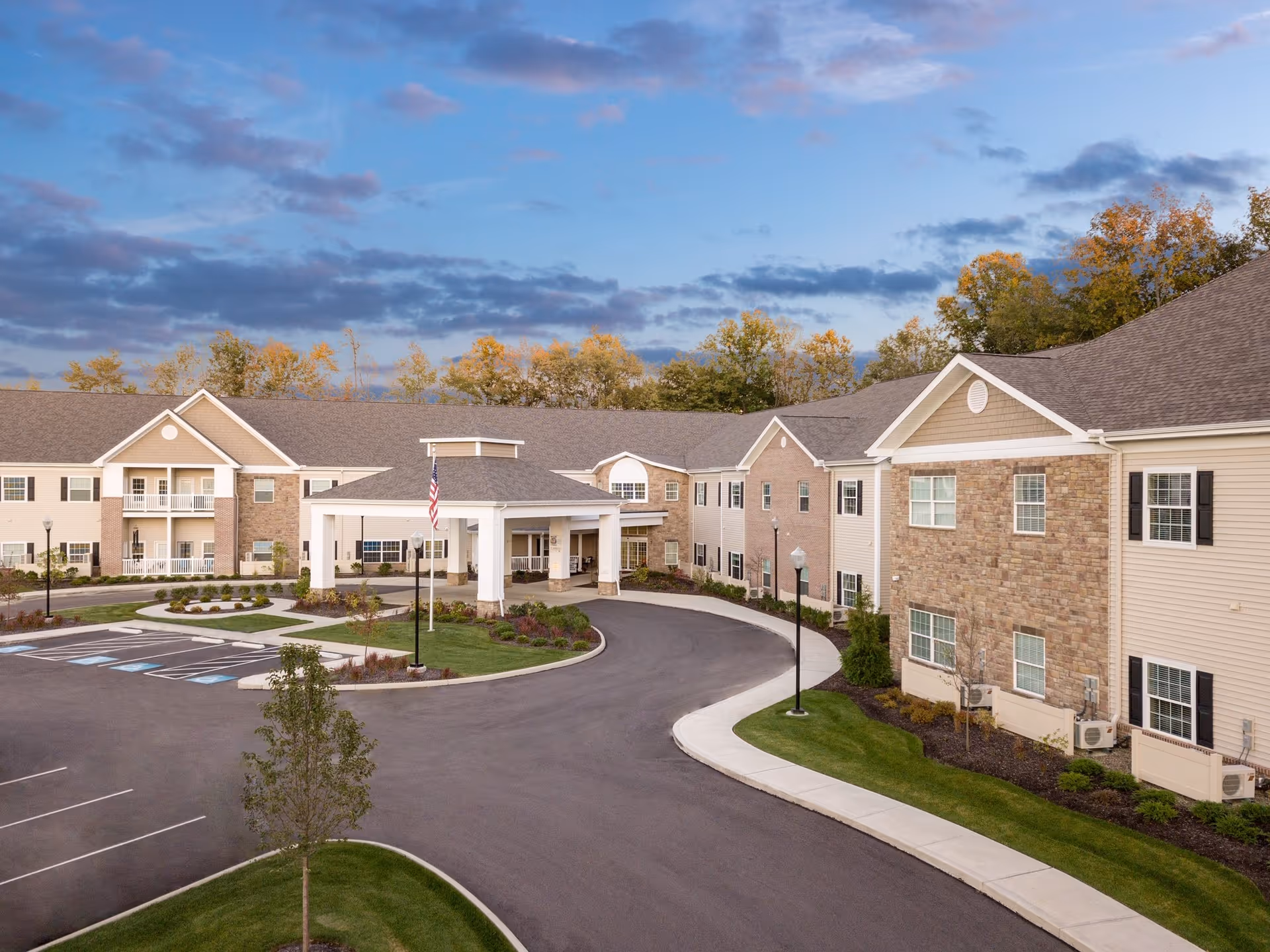 Exterior view of Danbury Brunswick senior living facility showing a large two-story building with beige siding and stone accents, a covered entrance with an American flag, a curved driveway, parking spaces including handicapped spots, and landscaped greenery under a partly cloudy sky.