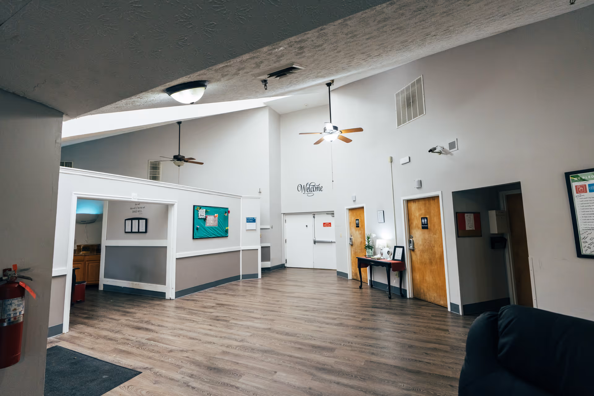 Spacious lobby of a senior living facility with a vaulted ceiling, ceiling fans, wood-look floors, a 'Welcome' sign, and doors with bulletin boards.