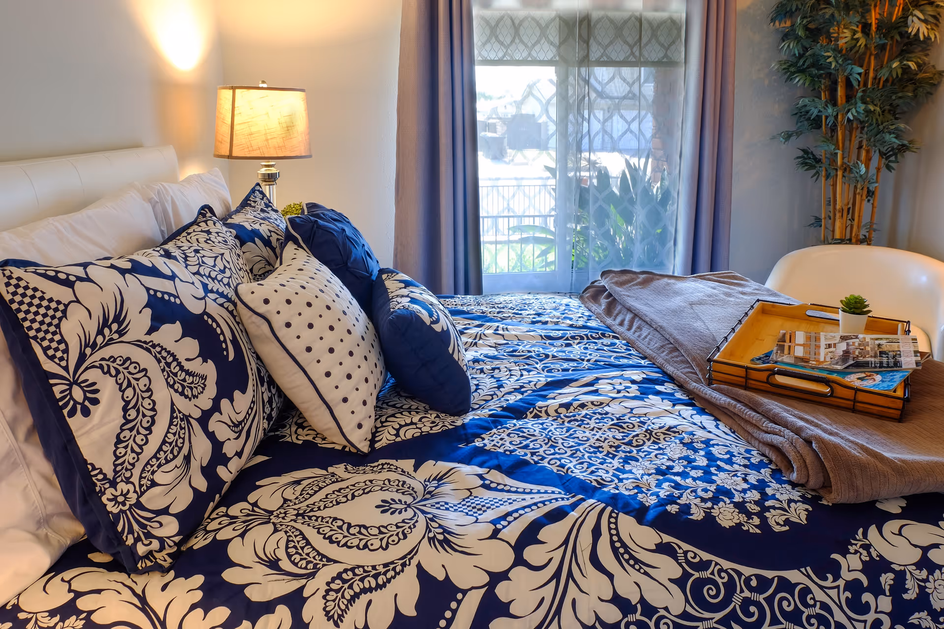 A neatly made bed with blue and white patterned bedding and multiple pillows in a bedroom. A bedside lamp is turned on, casting warm light on the wall. A window with sheer and solid curtains lets in natural light, and a small tray with magazines and a small plant rests on the bed. A tall potted plant is in the corner of the room.