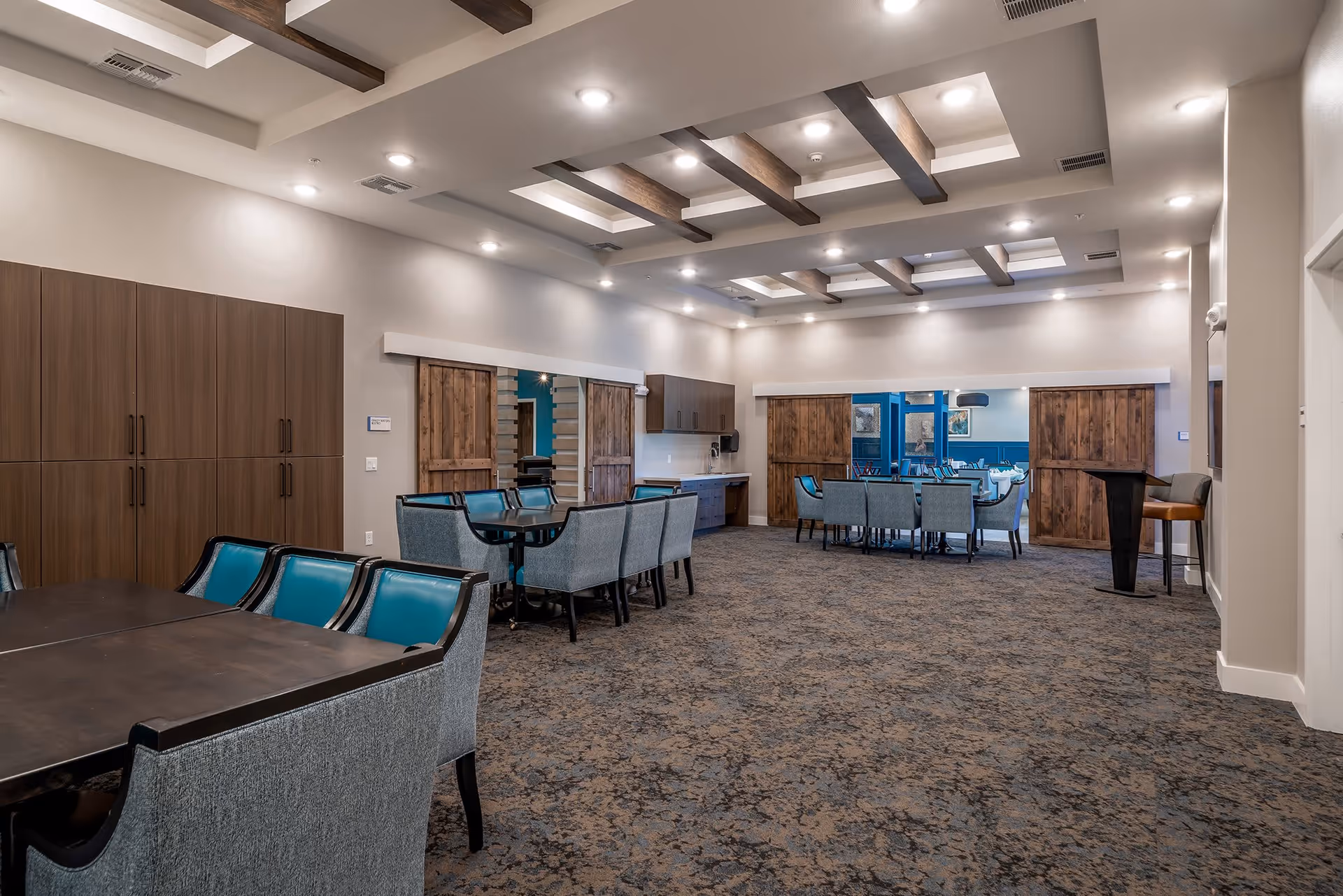 A spacious and well-lit interior room with multiple tables and chairs arranged for group seating. The ceiling features recessed lighting and wooden beams. There are wooden sliding barn doors on the far wall, with a glimpse into another room with additional seating. The room has a carpeted floor and neutral-colored walls with wooden cabinetry on one side.