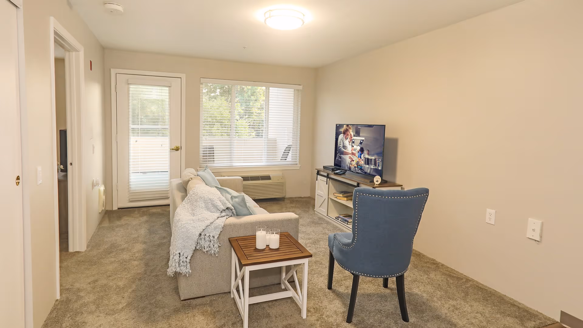 A cozy living room with beige walls and carpeted floor featuring a light gray sofa with a blue throw blanket and pillows, a small wooden side table with candles, a blue upholstered chair, and a TV on a white media console. There is a door and window with blinds letting in natural light.