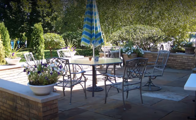Outdoor patio area with a glass-top table, metal chairs, a closed blue and green striped umbrella, and several potted plants on a stone floor surrounded by low brick walls and greenery.
