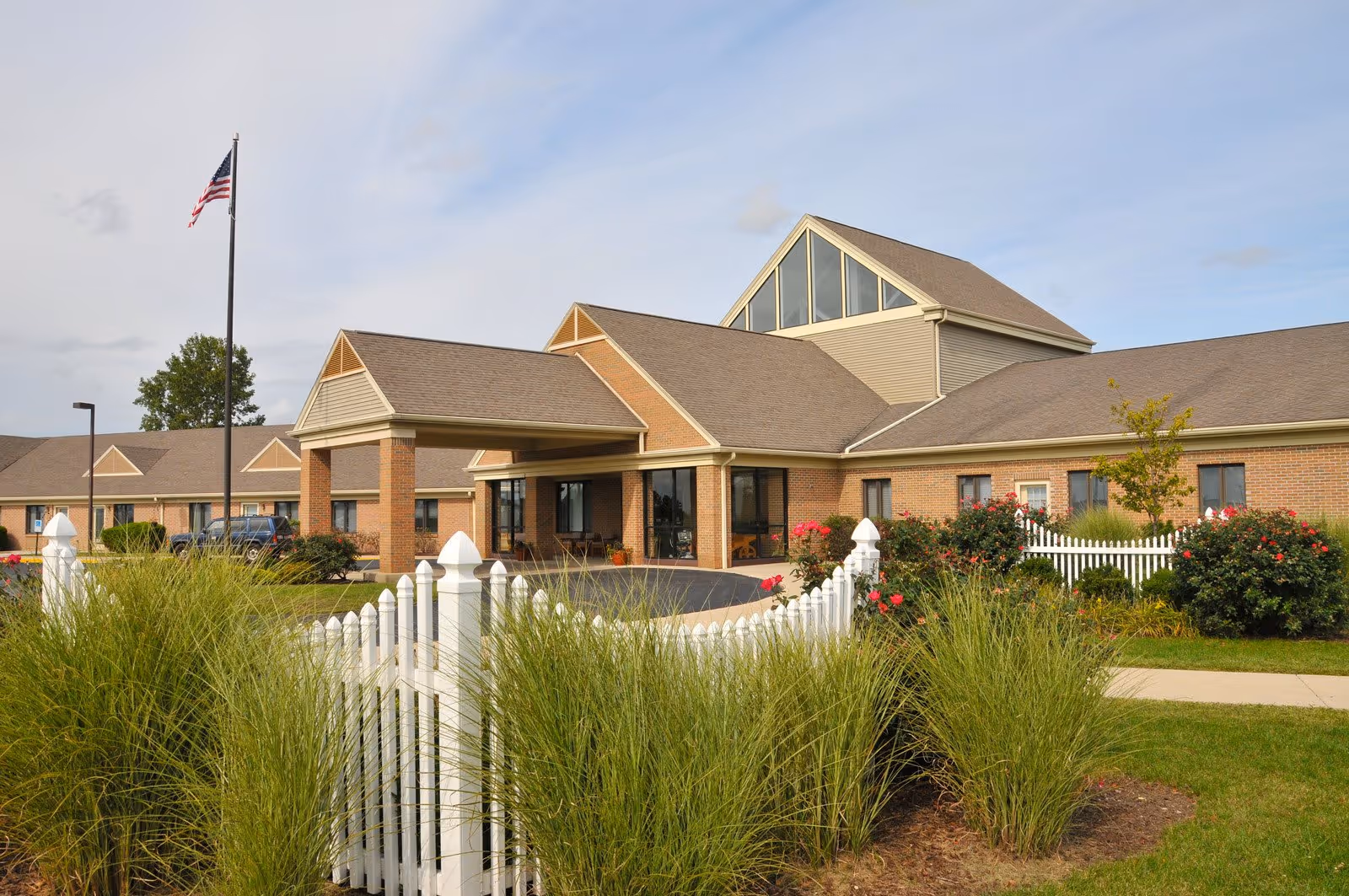 Front entrance of a brick senior living building with a white picket fence, landscaping and an American flag.