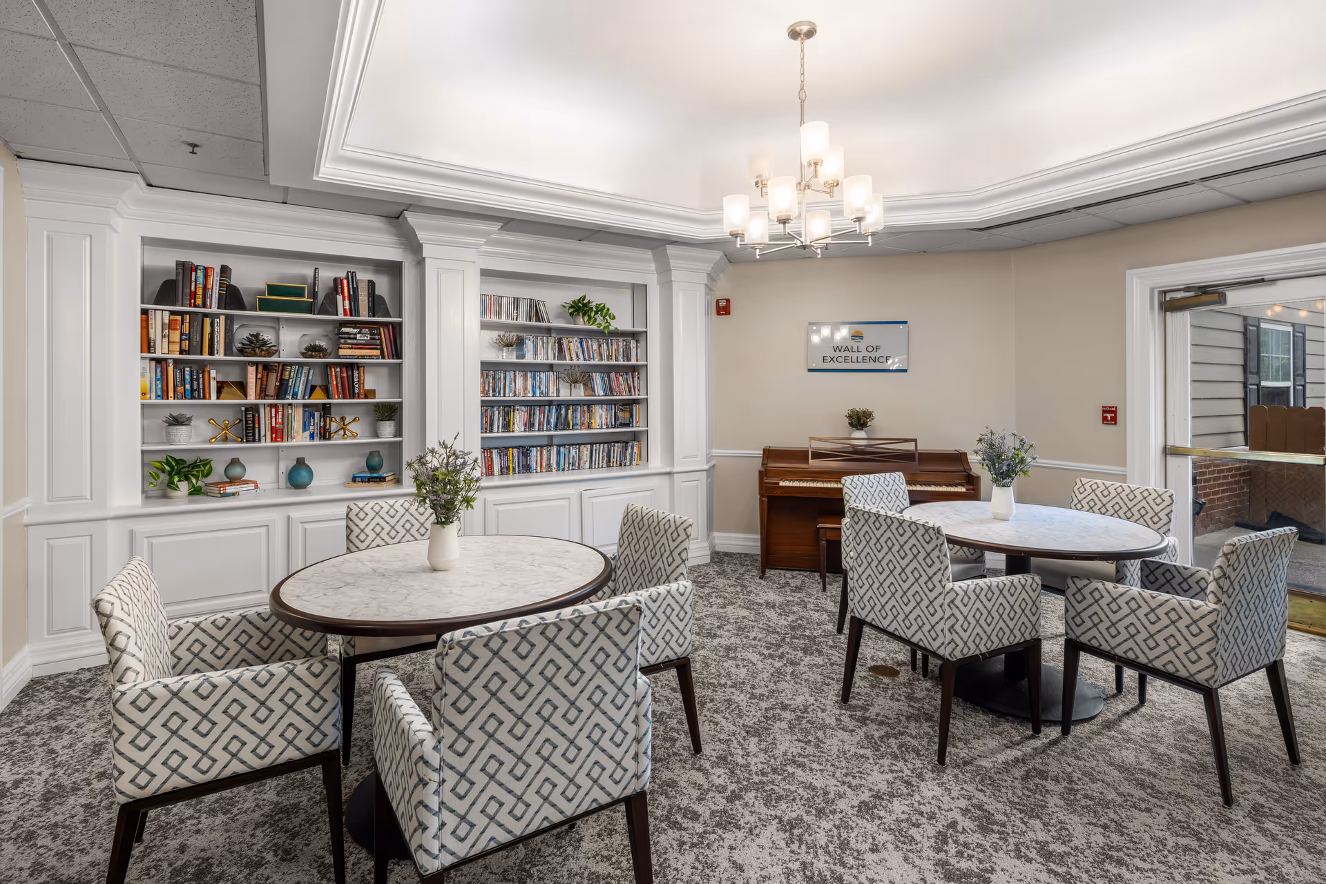 A cozy common area with two round marble tables surrounded by patterned armchairs. Behind the tables, there are built-in white shelves filled with books, DVDs, and decorative items. A wooden piano is placed against the wall under a sign that reads 'Wall of Excellence.' A chandelier with multiple lights hangs from the ceiling, and a glass door leads outside.