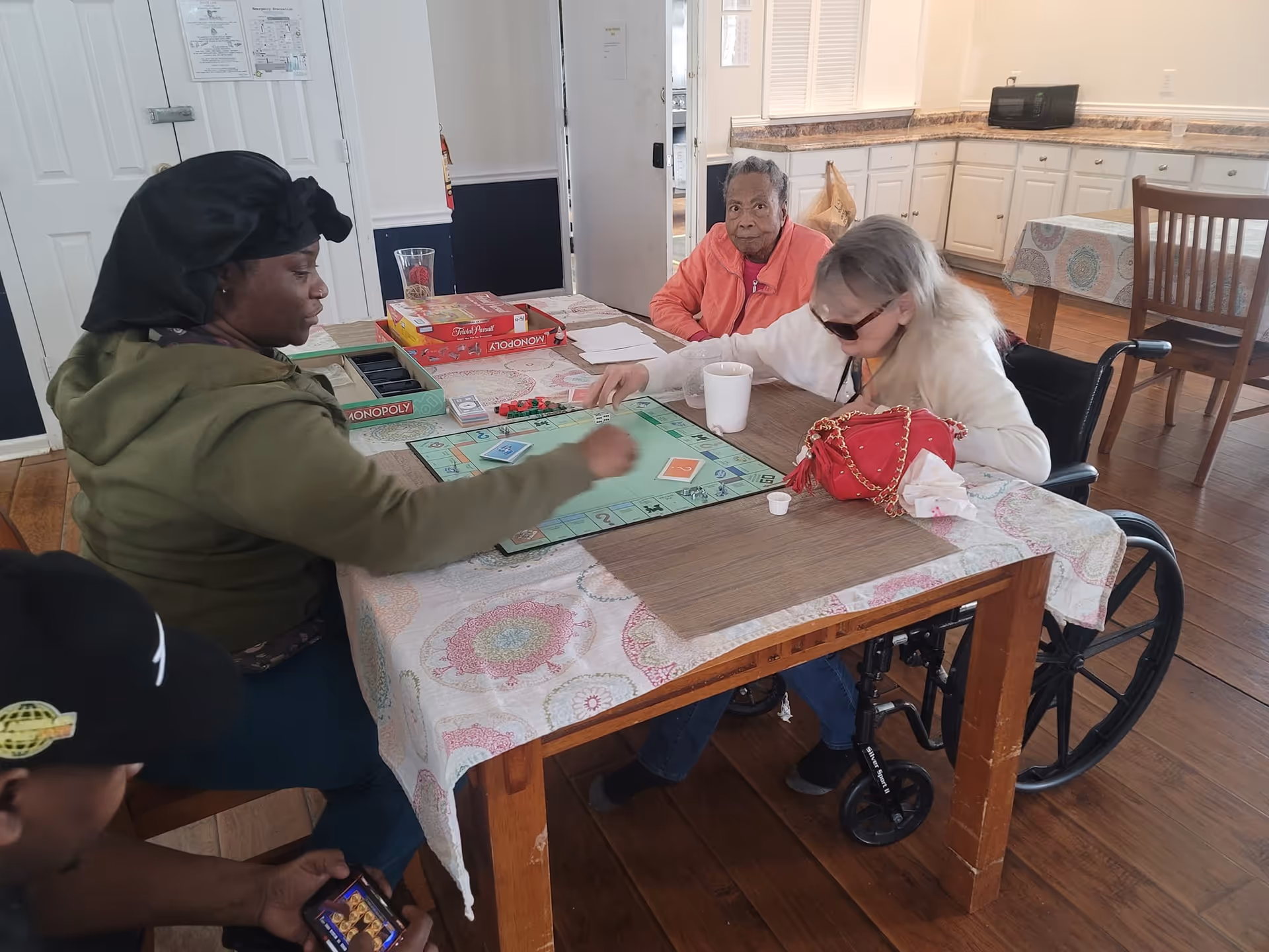 Three people sitting around a table playing a board game of Monopoly in a room with wooden floors and white cabinets in the background. One person is in a wheelchair, and there is a red purse on the table along with a white cup.