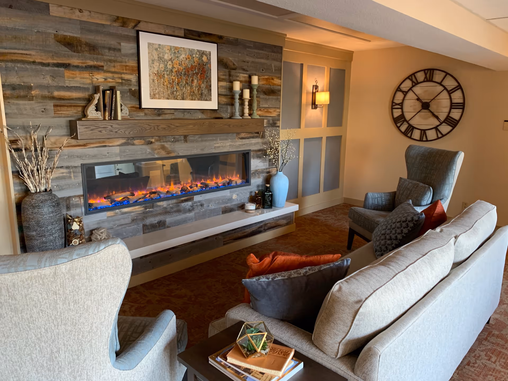 Cozy living room area with a modern electric fireplace set in a rustic wooden wall. The room features a beige sofa with multiple cushions, two upholstered armchairs, a large wall clock, and decorative items including a vase with branches and candles on a mantel. Warm lighting creates a comfortable atmosphere.