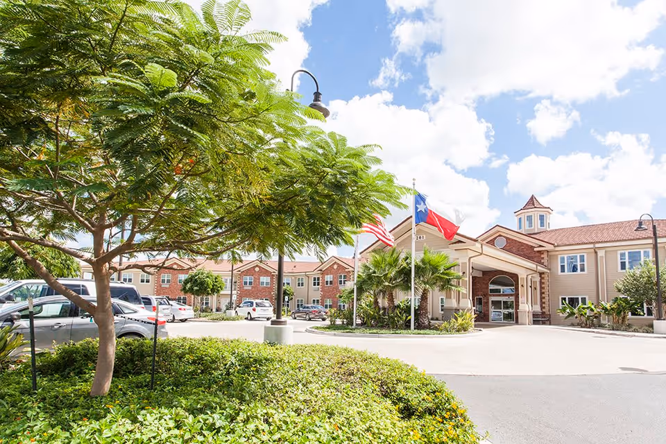Exterior view of The Gardens at Brook Ridge Assisted Living & Memory Care facility showing a two-story building with a covered entrance, American and Texas flags, a parking lot with several cars, and landscaped greenery including trees and bushes under a partly cloudy sky.