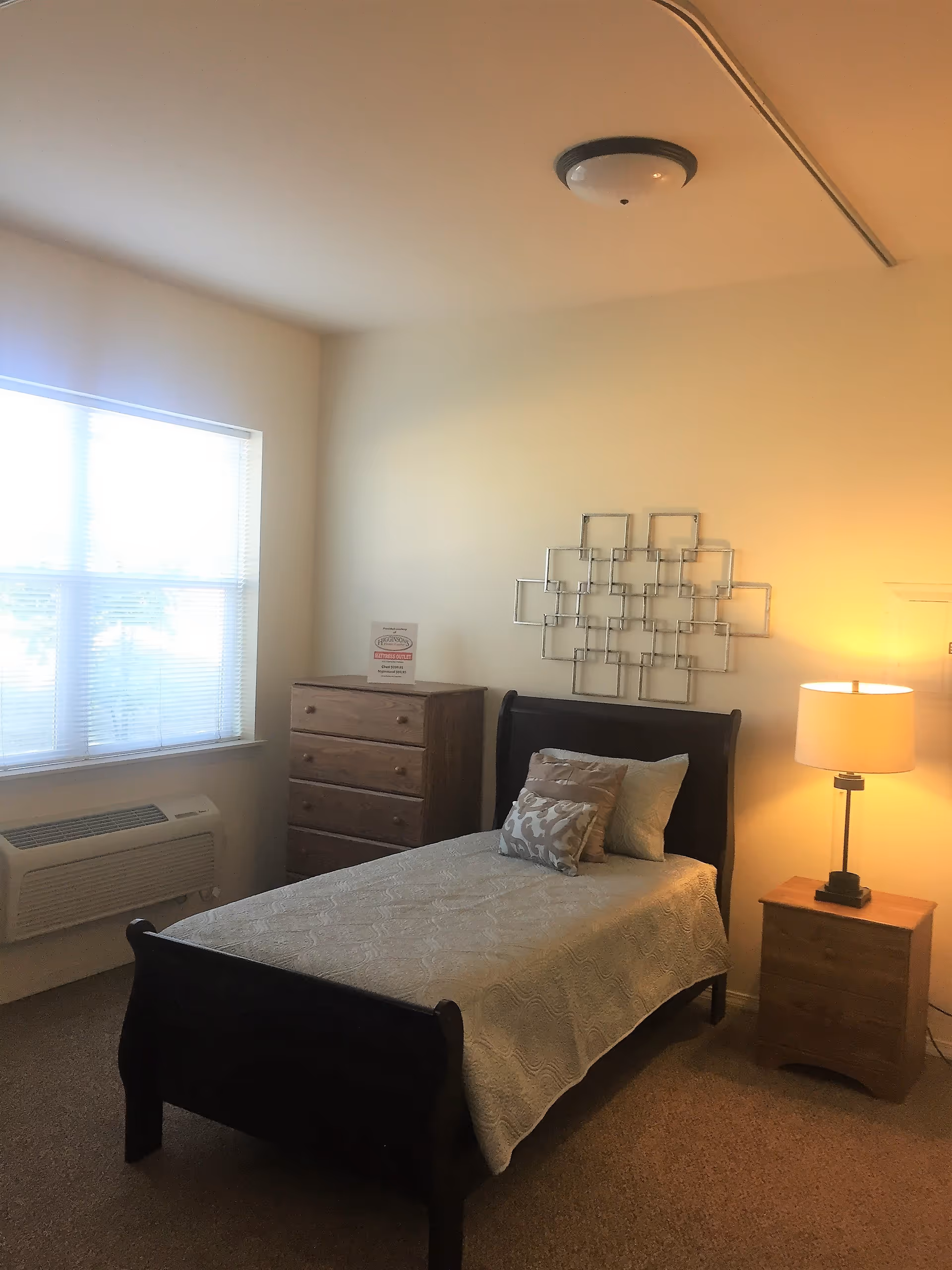 A simple bedroom with a single dark wooden bed covered with a light-colored quilt and three pillows. To the right of the bed is a wooden nightstand with a lamp emitting warm light. On the left side of the room is a wooden chest of drawers with a small sign on top. A large window with blinds allows natural light into the room. The walls are plain and light-colored, with a decorative metal wall art piece above the bed.