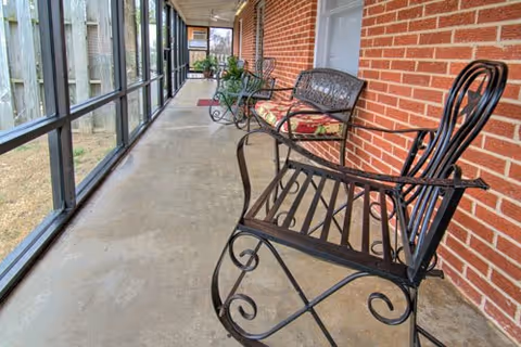 A long enclosed porch with a concrete floor and large windows on one side. The opposite wall is made of red brick. Several black metal benches with decorative designs and cushions are placed along the brick wall. There are some plants and a doormat visible further down the porch.