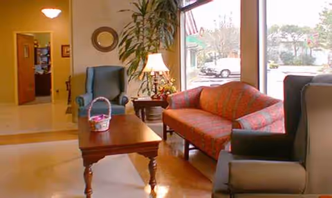Sunlit senior living lounge with colorful sofa, chairs, coffee table, lamp, and plants.