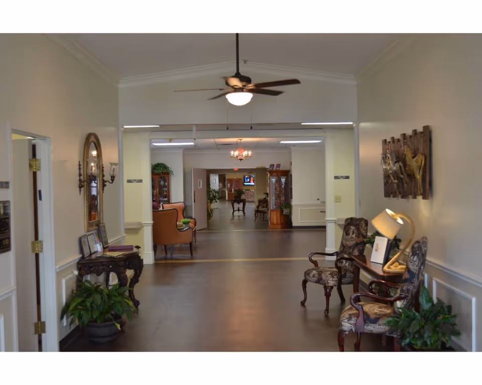 Interior hallway of a senior living facility with decorative chairs, a small table with a lamp, plants, a ceiling fan, and wall art. The hallway leads to a larger common area with more seating and a television in the background.
