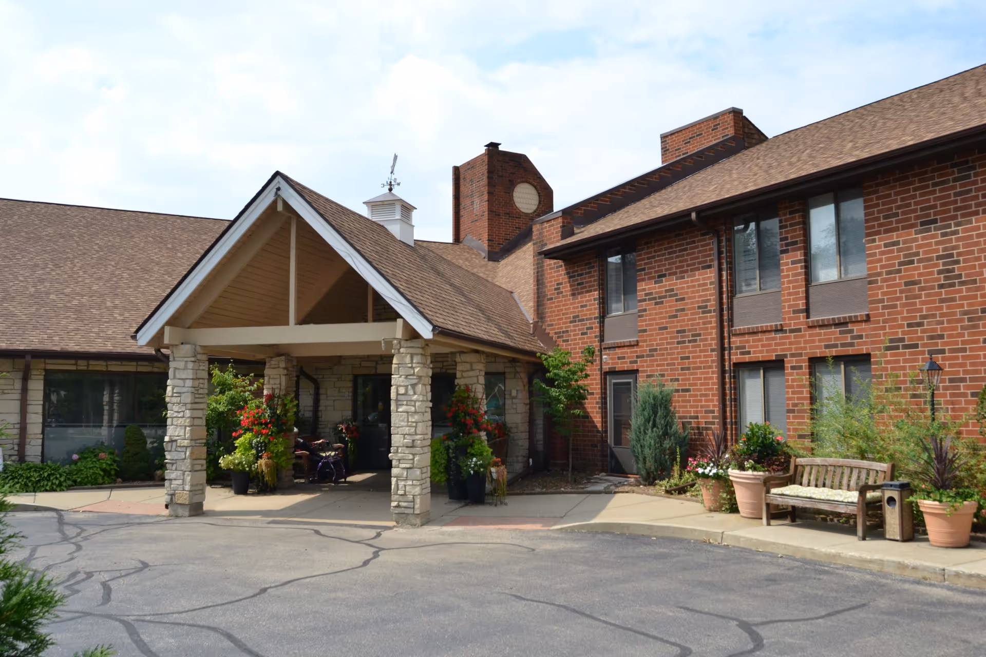 Exterior view of a senior living facility with a covered entrance supported by stone pillars. The building has a brick facade with multiple windows, a bench, and several potted plants near the entrance. The sky is partly cloudy.