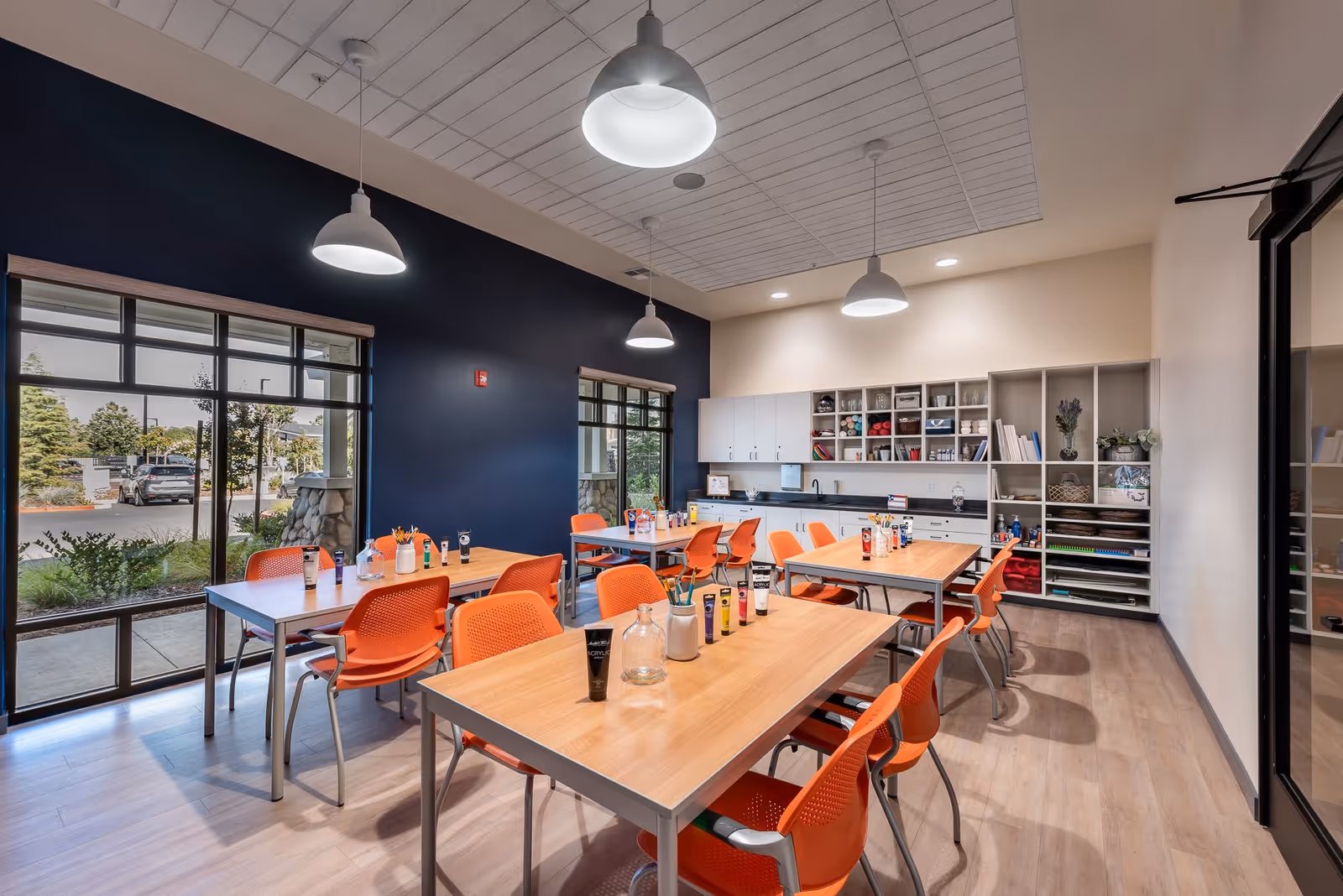 A bright and modern activity room with several wooden tables and orange chairs arranged neatly. The tables have various art supplies including paint tubes, brushes, and jars. Large windows on one side let in natural light and offer a view of the outdoors. The back wall features shelves and cabinets filled with more art materials and supplies. The room has a light wood floor and pendant lights hanging from the ceiling.