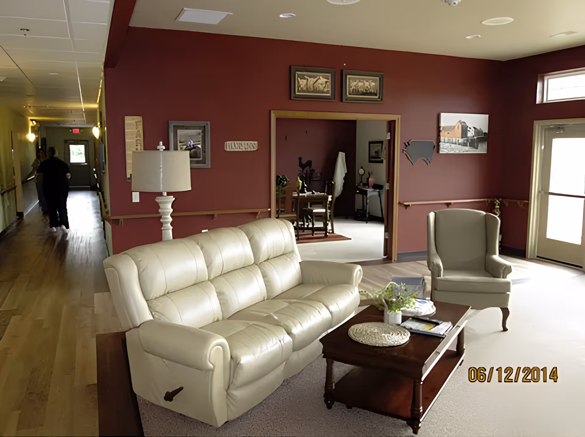 A cozy living room area with a cream-colored leather sofa and matching armchair. A wooden coffee table with a plant and some magazines sits in front of the sofa. The walls are painted a deep red, decorated with framed pictures and wall art. A hallway extends to the left with two people walking away, and a doorway in the center leads to another room with a wooden dining table and chairs. Natural light enters through a window and a glass door on the right.