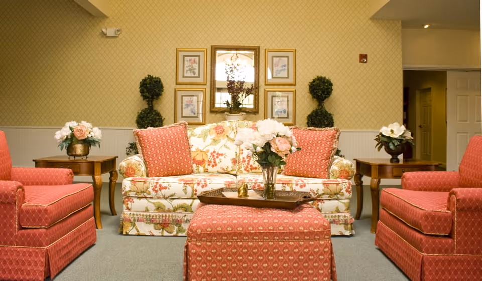 A cozy living room area with a floral patterned sofa adorned with two red patterned cushions, flanked by two matching red armchairs. In front of the sofa is a red patterned ottoman with a tray holding a vase of flowers. Two wooden side tables with flower arrangements sit beside the sofa. The wall behind features a mirror and framed floral artwork, with two topiary plants on either side.