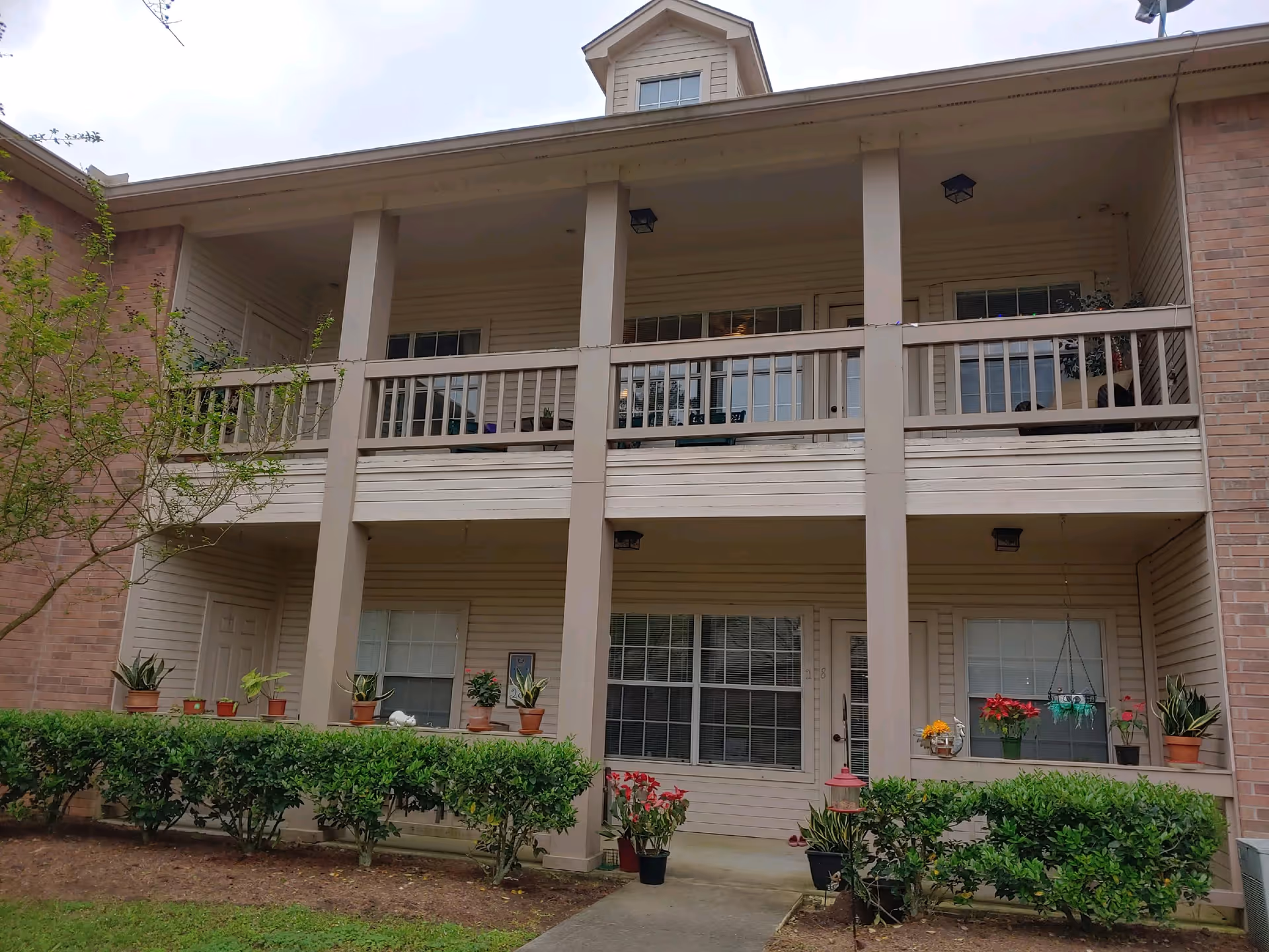 Exterior view of a two-story residential building with beige siding and brick accents. The building features a covered balcony on the second floor with railings and multiple windows and doors on both floors. There are various potted plants and flowers placed along the ground floor and balcony, with neatly trimmed bushes and a small tree in front of the building.