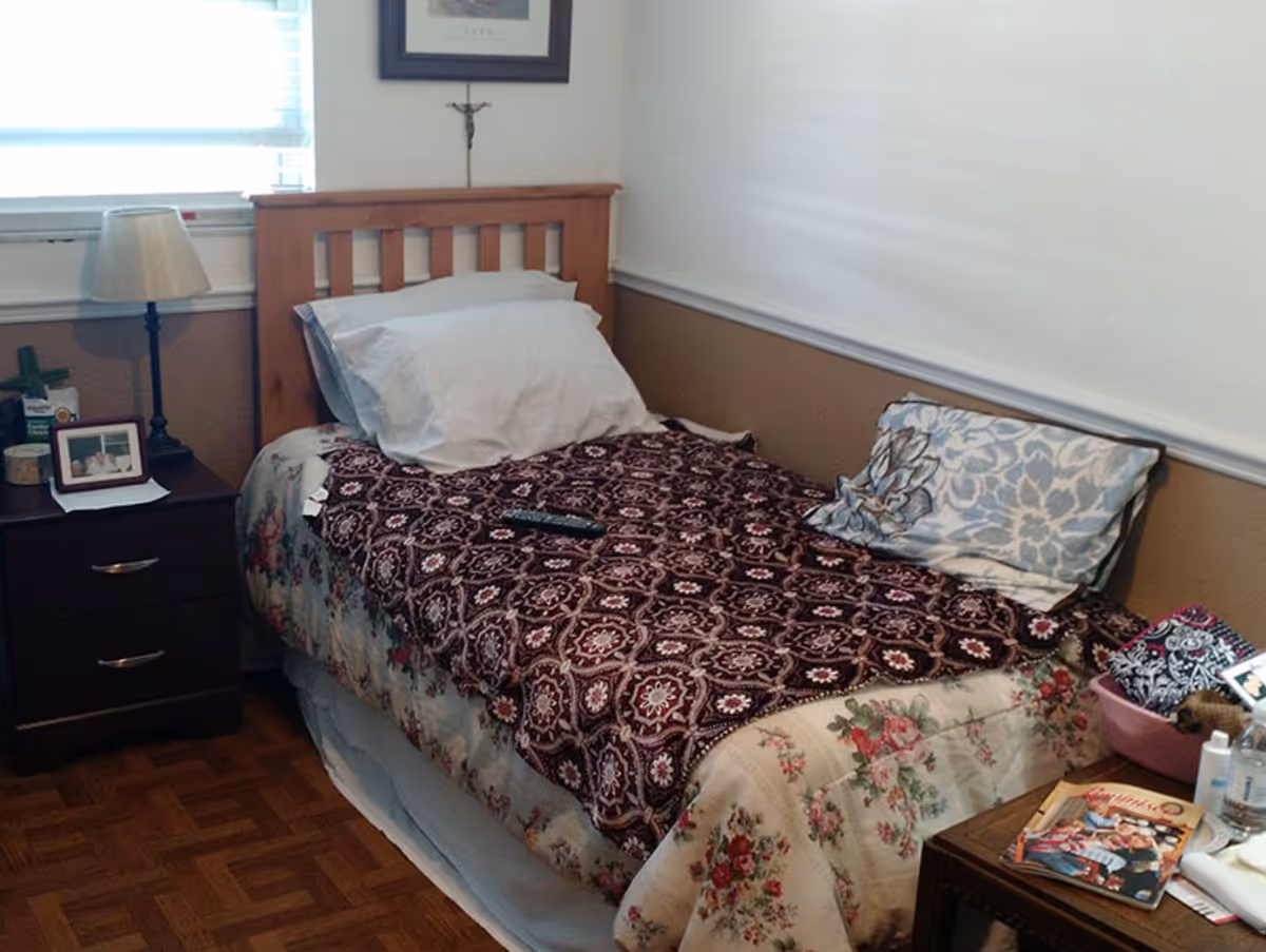 A single bed in a small bedroom with floral and patterned bedcovers, pillows, a wooden headboard, bedside tables, and a lamp.
