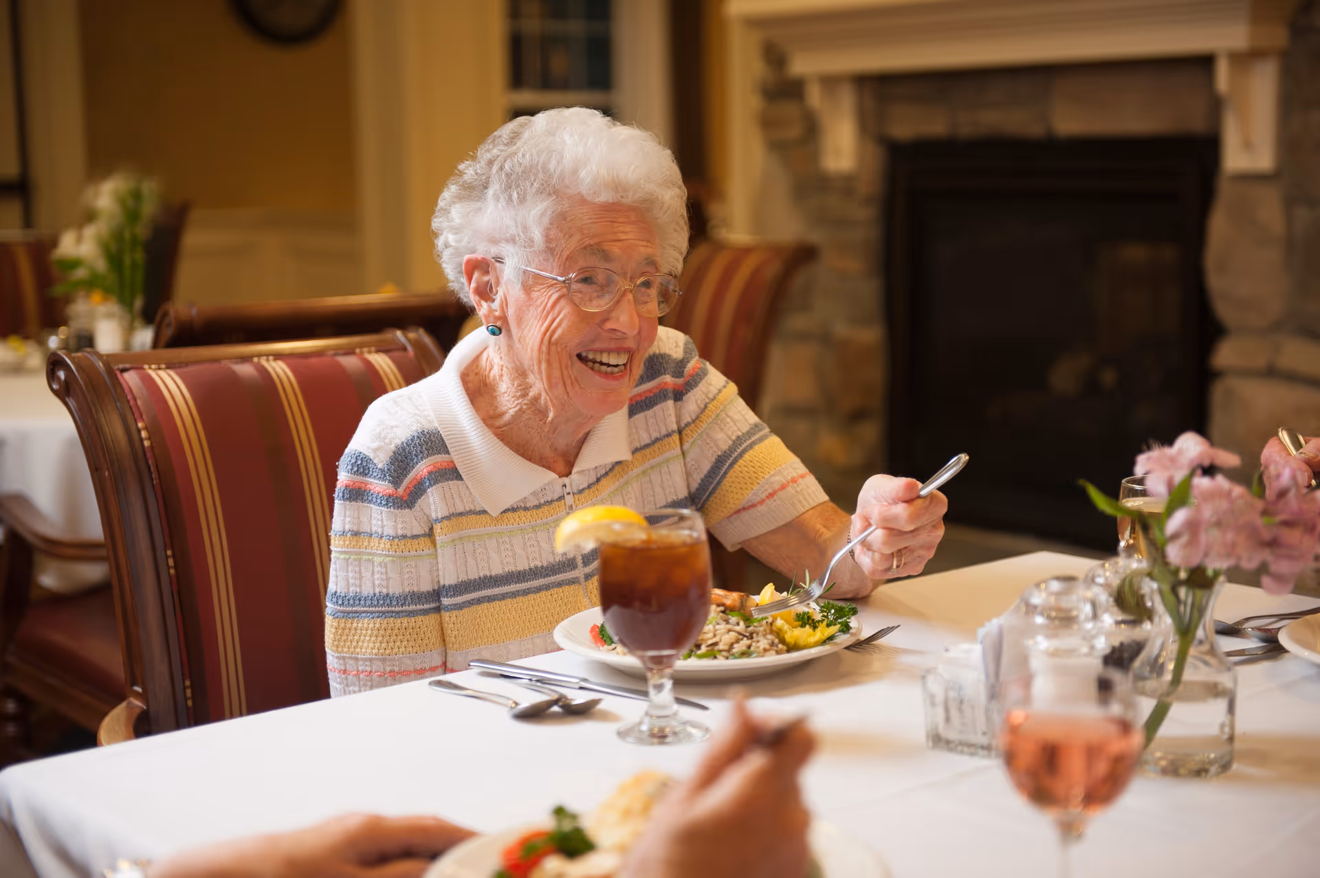 An elderly woman smiling while eating a meal at a dining table with place settings and a floral centerpiece.