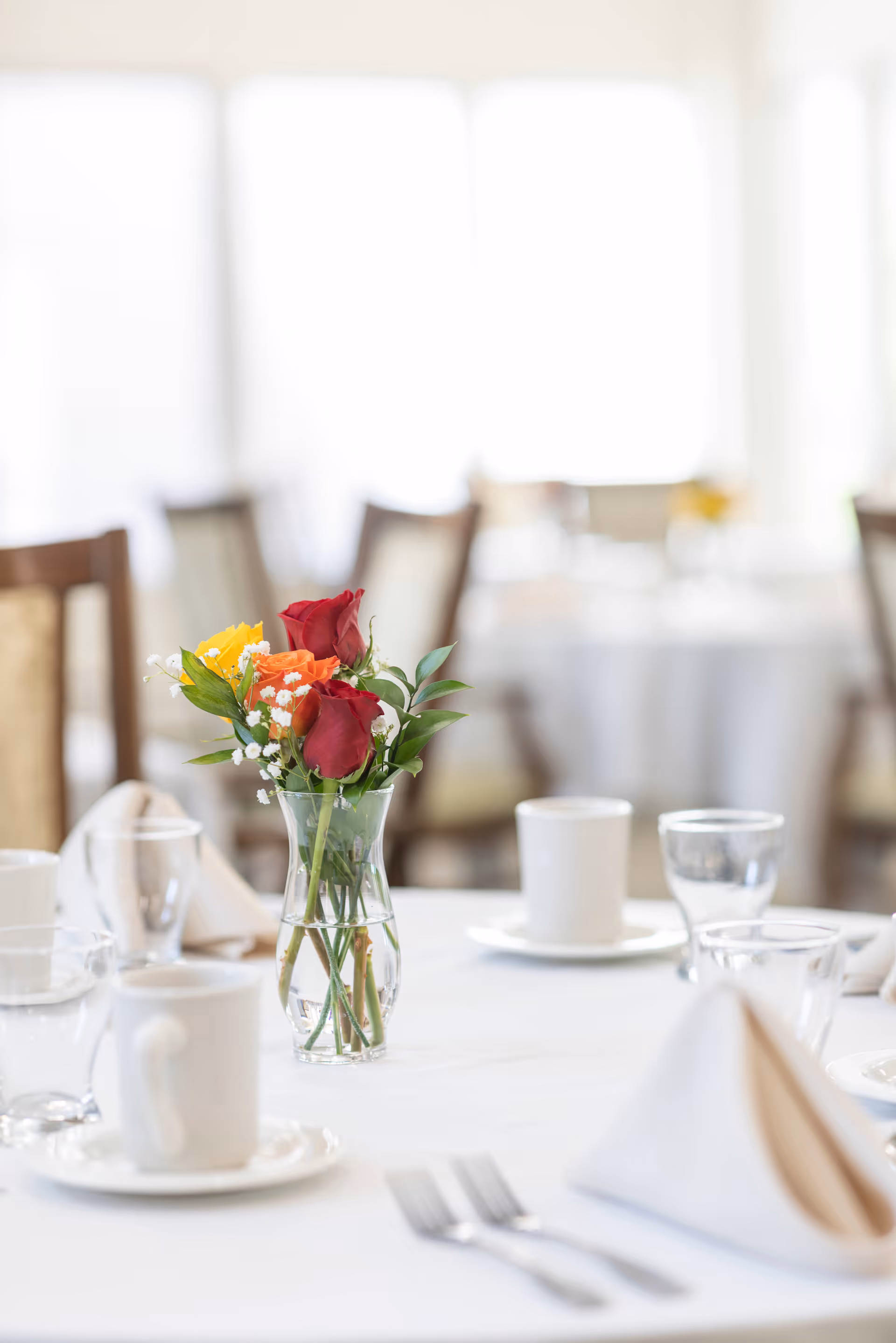 A dining table set with white tablecloth, white coffee cups on saucers, clear drinking glasses, folded beige napkins, and a small glass vase with a bouquet of red, orange, and yellow roses with greenery in the center.