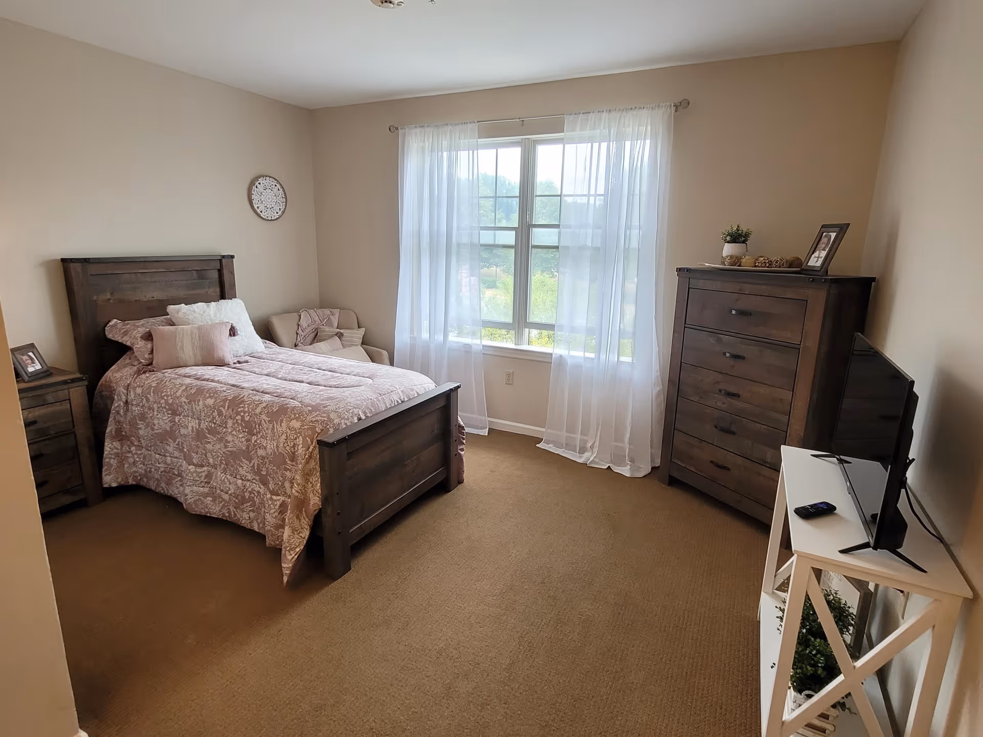 A cozy bedroom with a wooden bed frame and matching nightstand and dresser. The bed is made with a pink and white floral comforter and several pillows. There is a window with sheer white curtains letting in natural light, a small armchair in the corner, and a TV on a white stand opposite the bed.