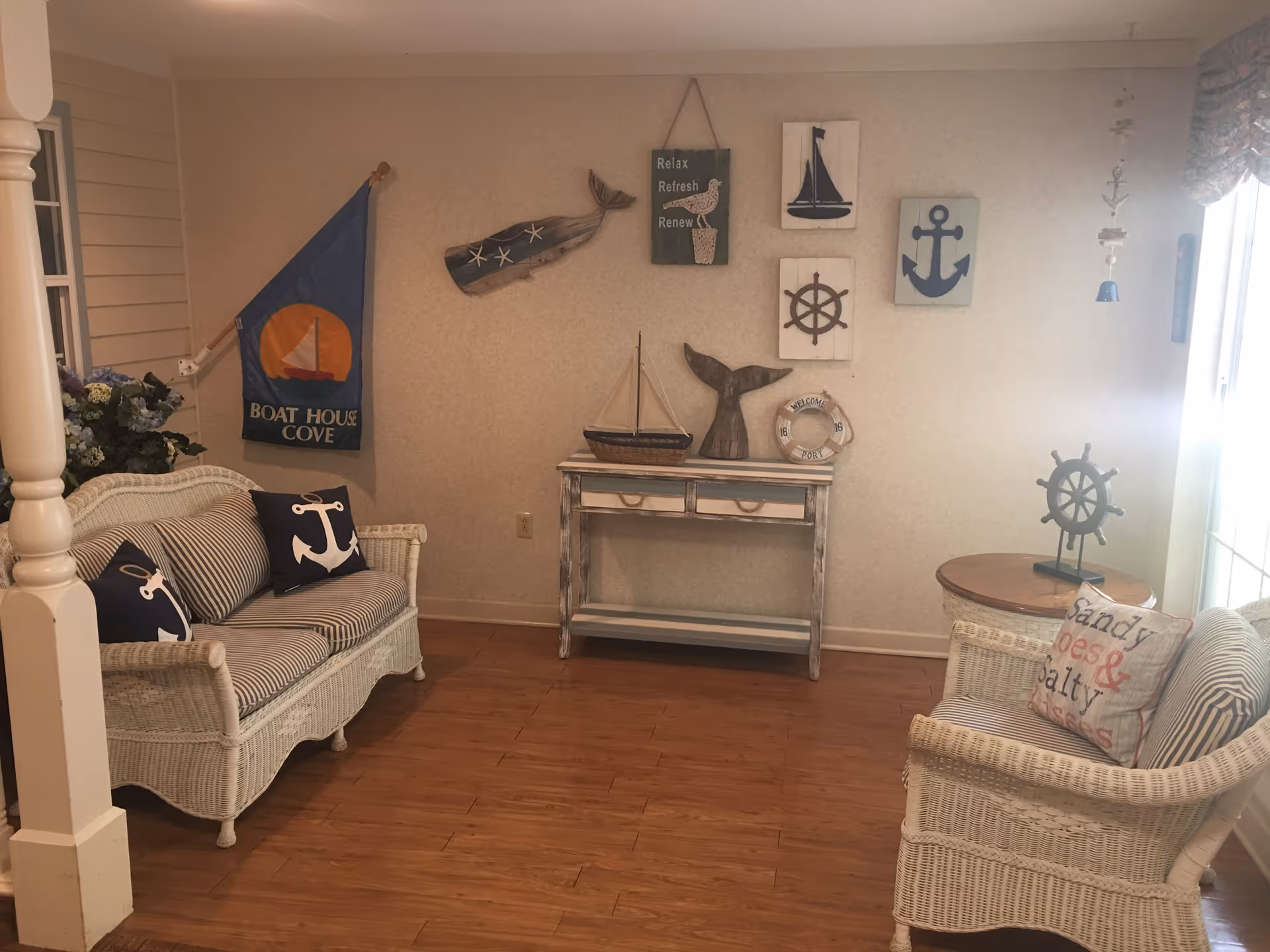 A cozy sitting area with nautical-themed decor including a wicker loveseat and chair with anchor and striped cushions, a small wooden table, and a console table against the wall. The wall is decorated with various nautical items such as a whale, sailboat, ship wheel, anchor, and a life preserver. A flag with the text 'Boat House Cove' hangs on the left wall. The floor is wooden, and sunlight streams in from a window on the right.