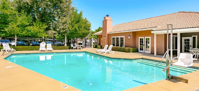 Outdoor swimming pool area at Creekside Village Apts with lounge chairs around the pool, a building with a tiled roof and large windows, and trees in the background under a clear blue sky.