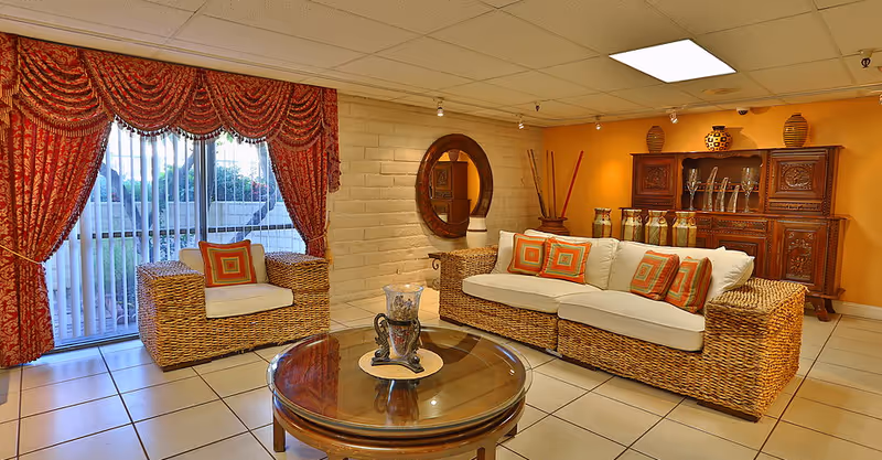 A cozy living room with wicker furniture including a sofa and an armchair, both with white cushions and orange patterned throw pillows. There is a round glass-top coffee table in the center with a decorative candle holder. The room has tiled flooring, a large window with red patterned curtains, a round wooden mirror on a white brick wall, and an ornate wooden cabinet against an orange wall with decorative vases on top.