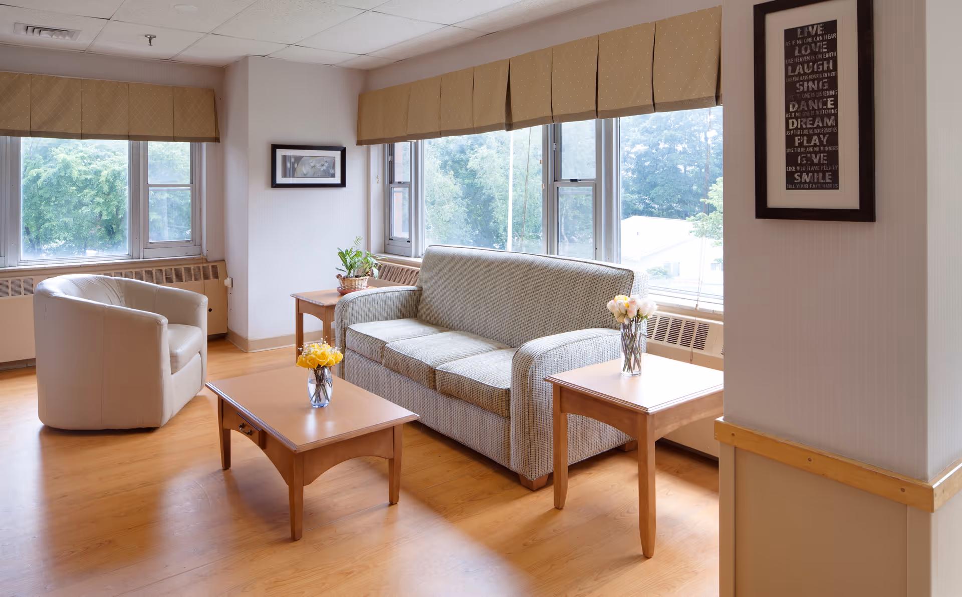 A bright and cozy living room area with large windows letting in natural light. The room features a beige armchair, a striped sofa, two wooden side tables with flower vases, and a wooden coffee table with a flower vase. The walls are light-colored with framed artwork and a motivational quote. The floor is wooden.