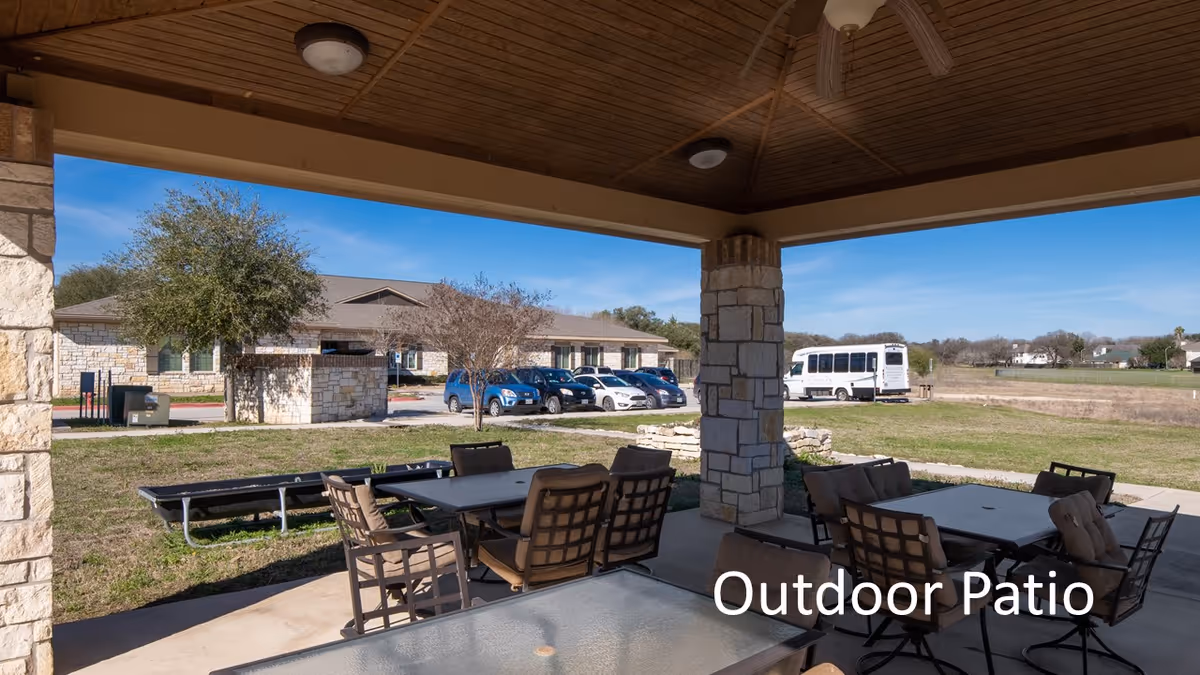 Covered outdoor patio area with cushioned chairs and tables, stone pillars, and a wooden ceiling. Beyond the patio, there is a grassy area, a parking lot with several cars, a white shuttle bus, and a single-story stone building under a clear blue sky.