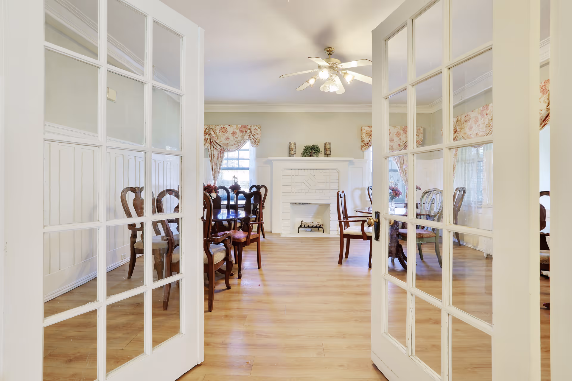 Bright dining room seen through glass-paneled double doors with wooden tables and chairs, a white decorative fireplace and ceiling fan.