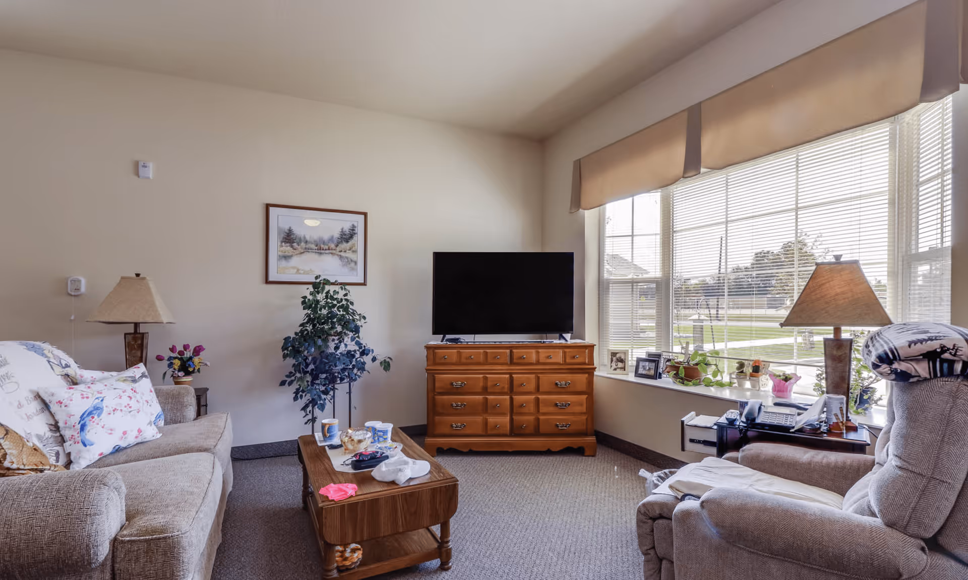 A cozy living room in Arbor Grove Assisted Living & Memory Care featuring a beige sofa with floral pillows, a wooden coffee table with cups and small items, a wooden dresser with a flat-screen TV on top, a large window with blinds and a beige valance letting in natural light, a recliner chair with a blanket, two table lamps, a potted plant, and framed pictures on the window sill.