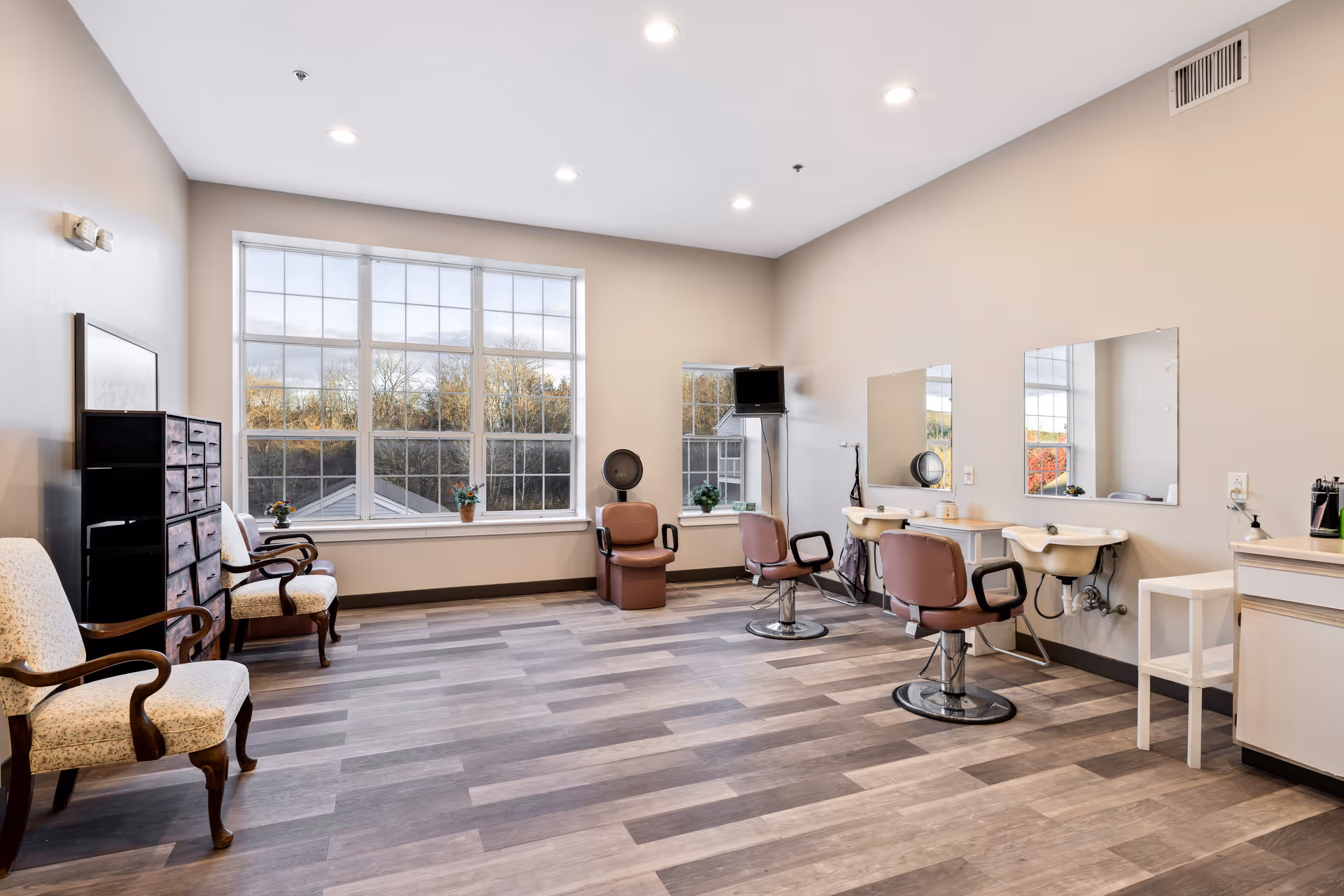 A bright and spacious salon area in a senior living community with large windows letting in natural light. The room features two salon chairs in front of mirrors with sinks, two vintage-style armchairs, a small cabinet with drawers, and a wall-mounted TV. The floor is covered with wood-patterned vinyl planks, and the walls are painted a soft beige color.