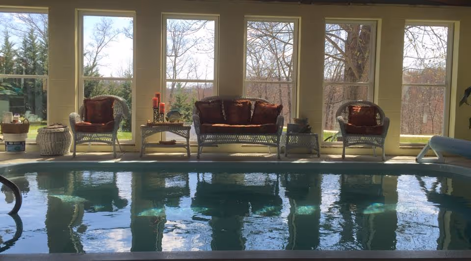 Indoor pool area with wicker chairs, large windows, and reflections on water.
