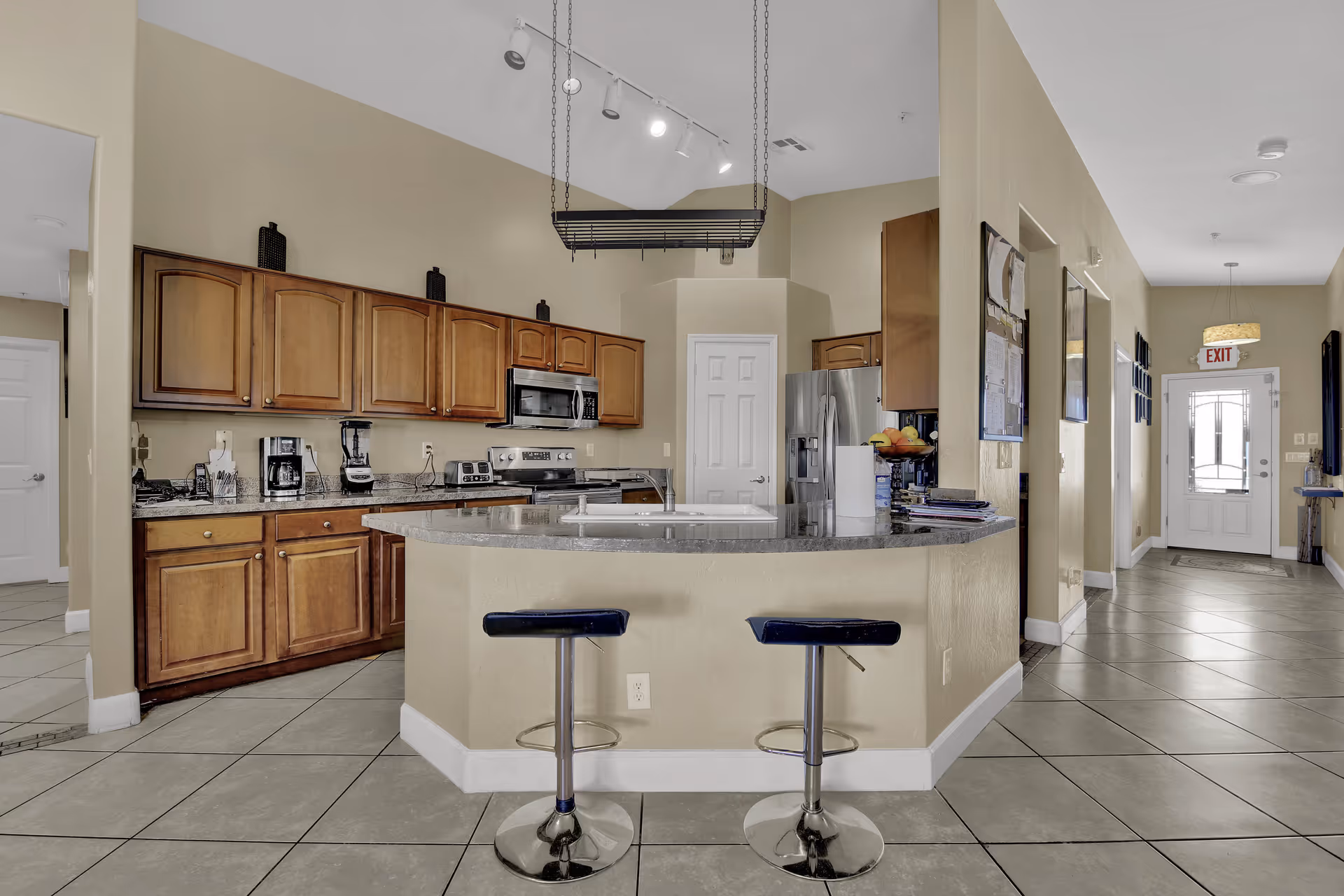 Open communal kitchen with a curved island and two bar stools, wooden cabinets, stainless steel appliances and a hallway leading to an exit door.
