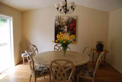 A round dining table with six chairs and a vase of flowers in a bright room.