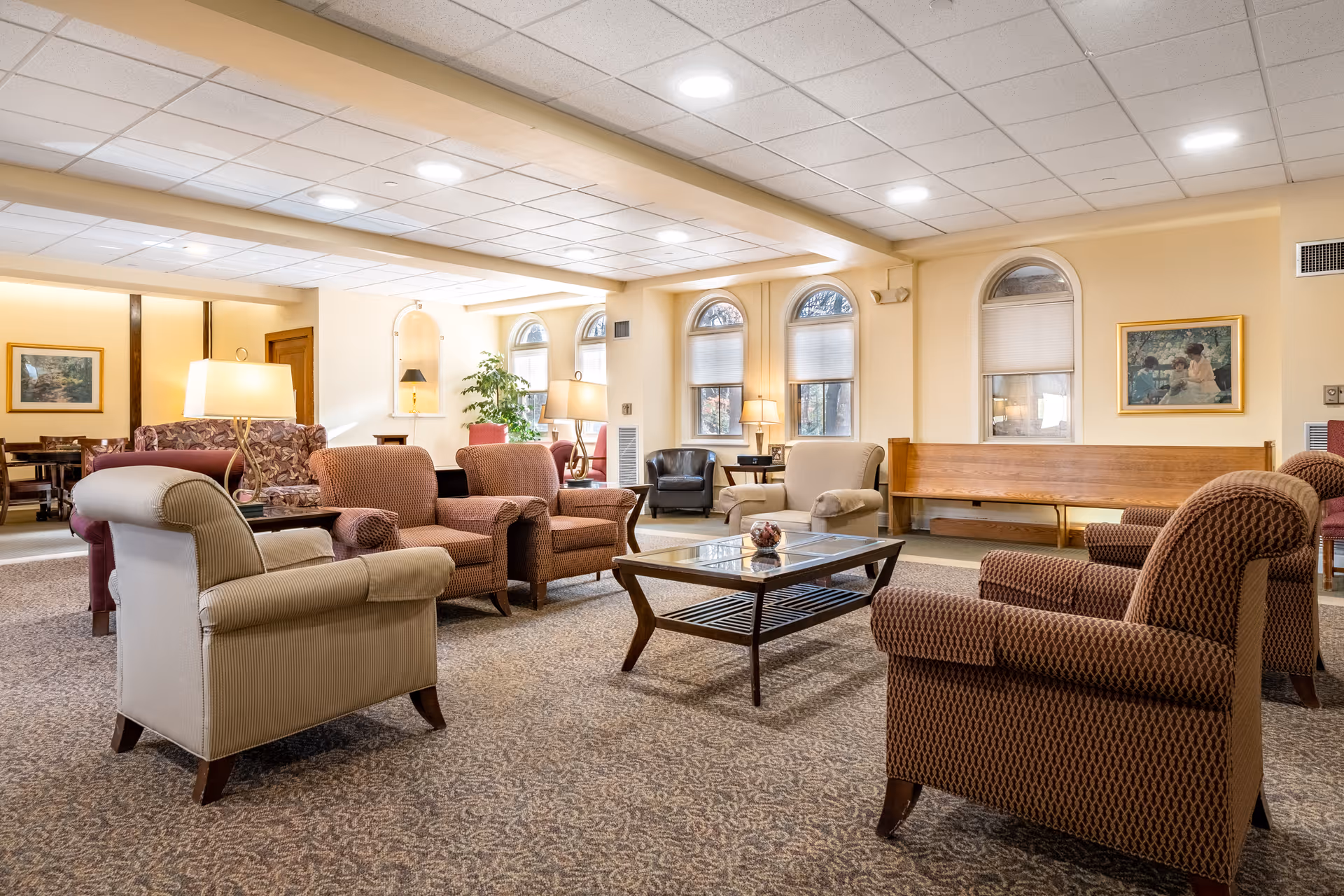 A spacious senior living facility common area with multiple upholstered armchairs and sofas arranged around a wooden coffee table. The room features large arched windows with blinds, framed artwork on the walls, table lamps, and a patterned carpet. The lighting is bright and warm, creating a welcoming atmosphere.