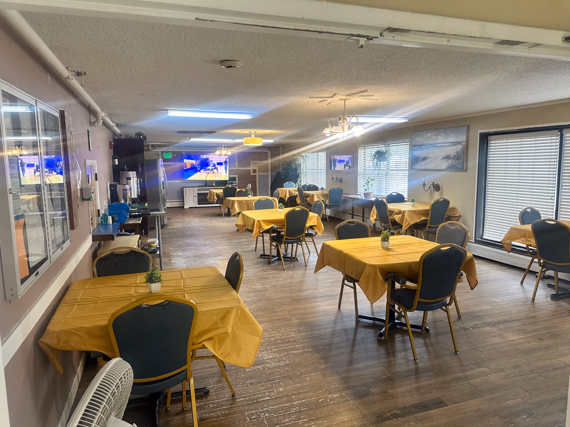 A dining room with several tables covered in yellow tablecloths, each surrounded by green cushioned chairs. The room has wooden flooring, large windows with blinds, and wall-mounted paintings. There are multiple light fixtures on the ceiling, and a television is visible on the far wall.
