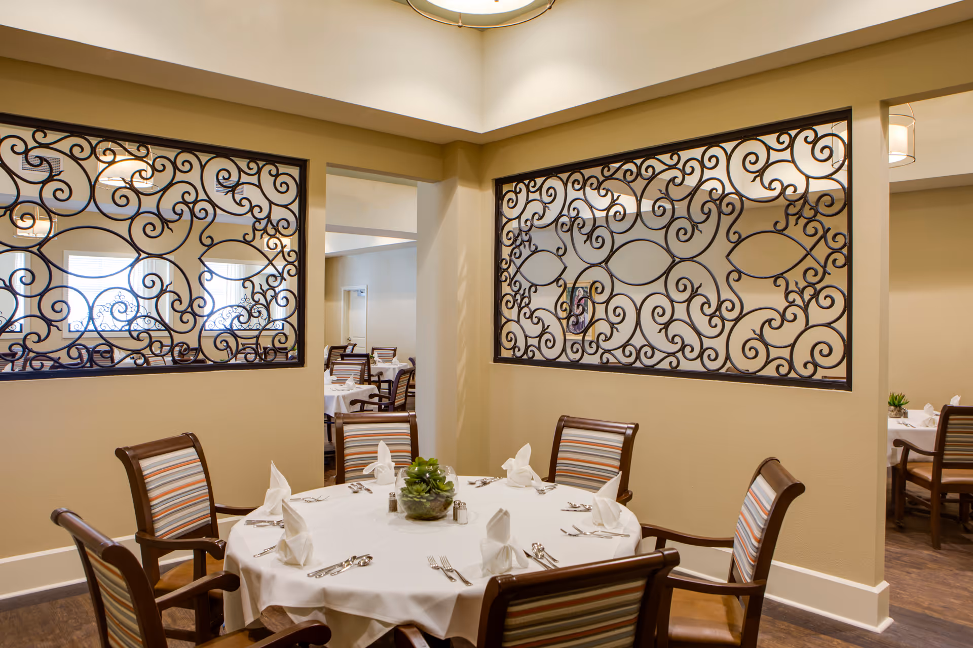 A dining area with a round table covered with a white tablecloth set for six people. Each place setting has a folded white napkin, silverware, and a small centerpiece with green plants. The room features beige walls with decorative wrought iron panels and wooden chairs with striped cushions.