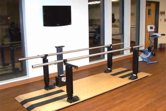 Indoor therapy gym area with parallel walking bars on a wooden platform, a wall-mounted TV, and an exercise bike.