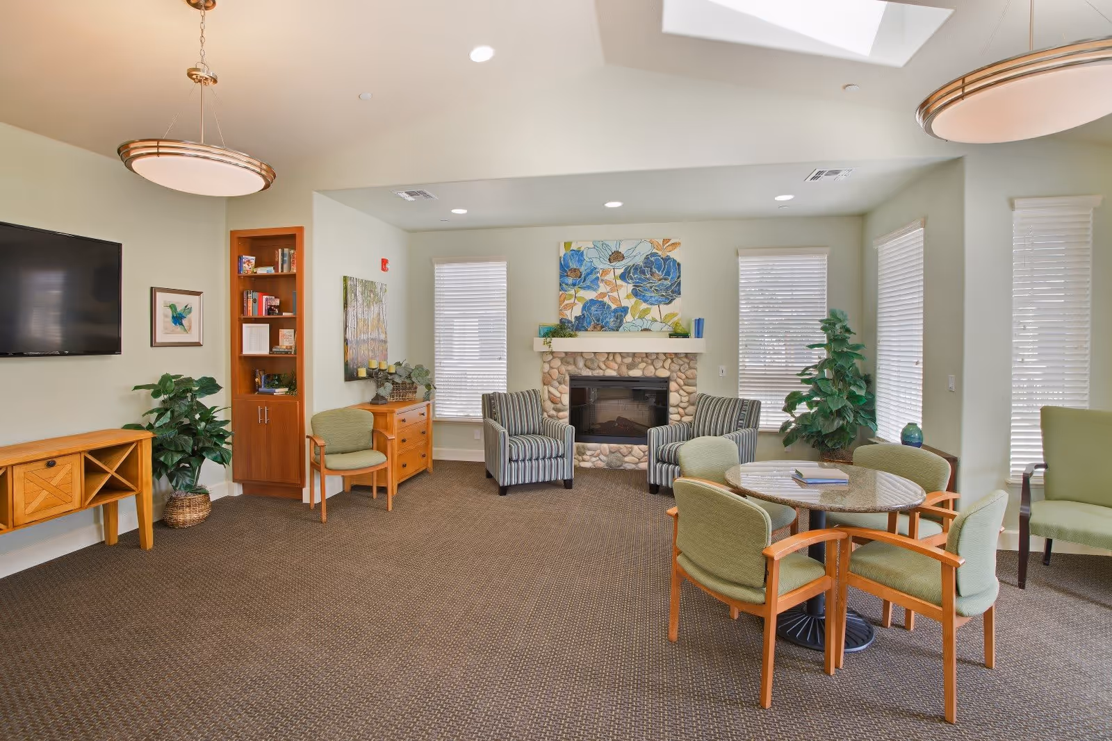 Communal living room with a stone fireplace, striped armchairs, a round table with green chairs, wall-mounted TV, and potted plants.