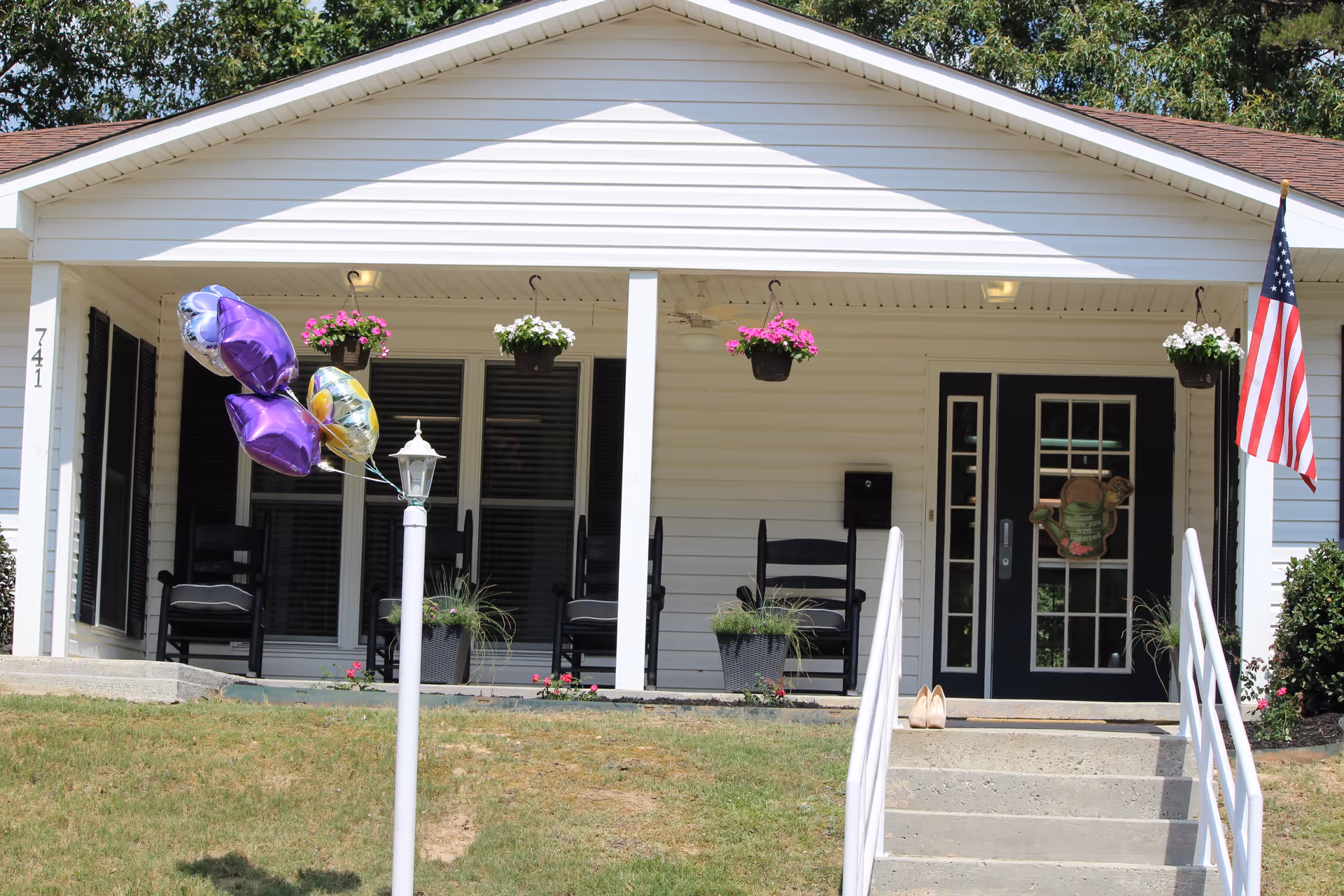 Front porch of a white single-story building with black-framed glass doors and windows. The porch has two black chairs, hanging flower pots with pink and white flowers, a white lamp post with purple and yellow balloons tied to it, and an American flag on the right side. Concrete steps with white railings lead up to the porch.