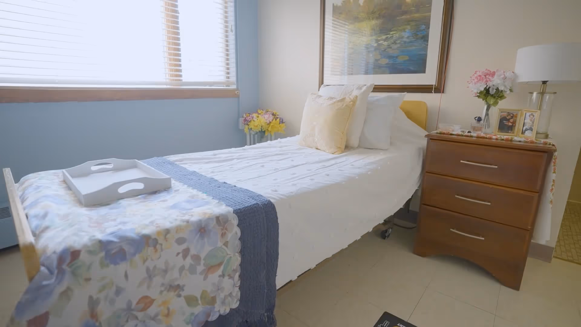 A neatly made single bed with white bedding and a blue throw blanket in a bright room with a window covered by blinds. A wooden nightstand with three drawers is beside the bed, holding a lamp, flowers, and framed photos. A floral table runner and a white tray are placed on the footboard of the bed.