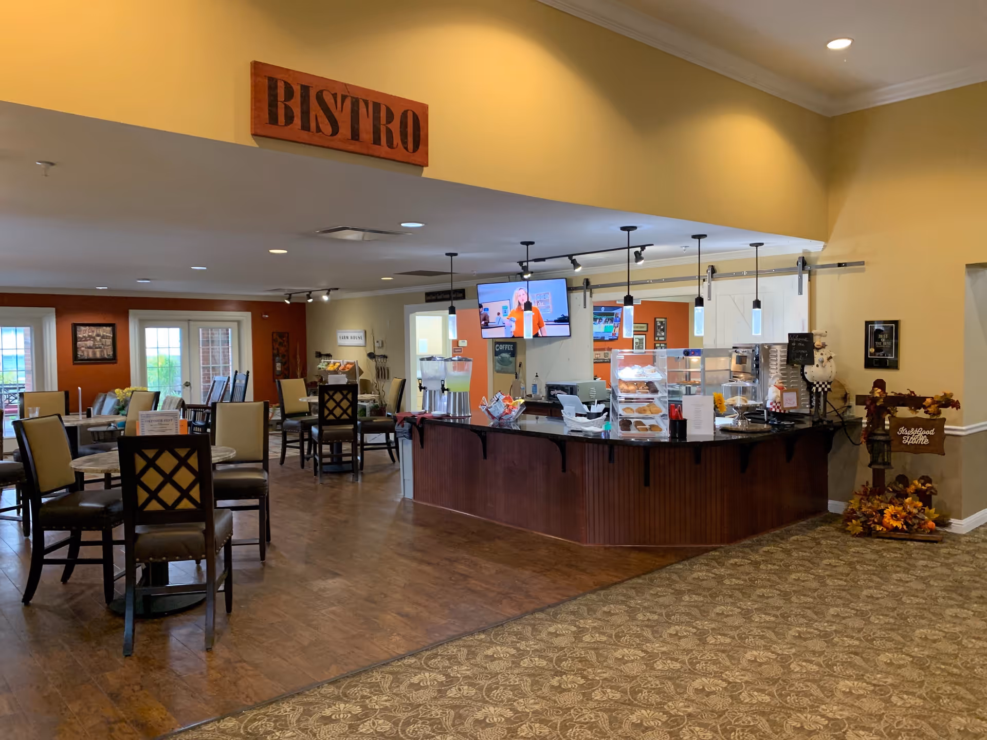 Interior view of a bistro area in a senior living facility with several tables and chairs arranged for dining. A counter displays baked goods and beverages, with a TV mounted on the wall behind it. The space has warm lighting and a welcoming atmosphere.