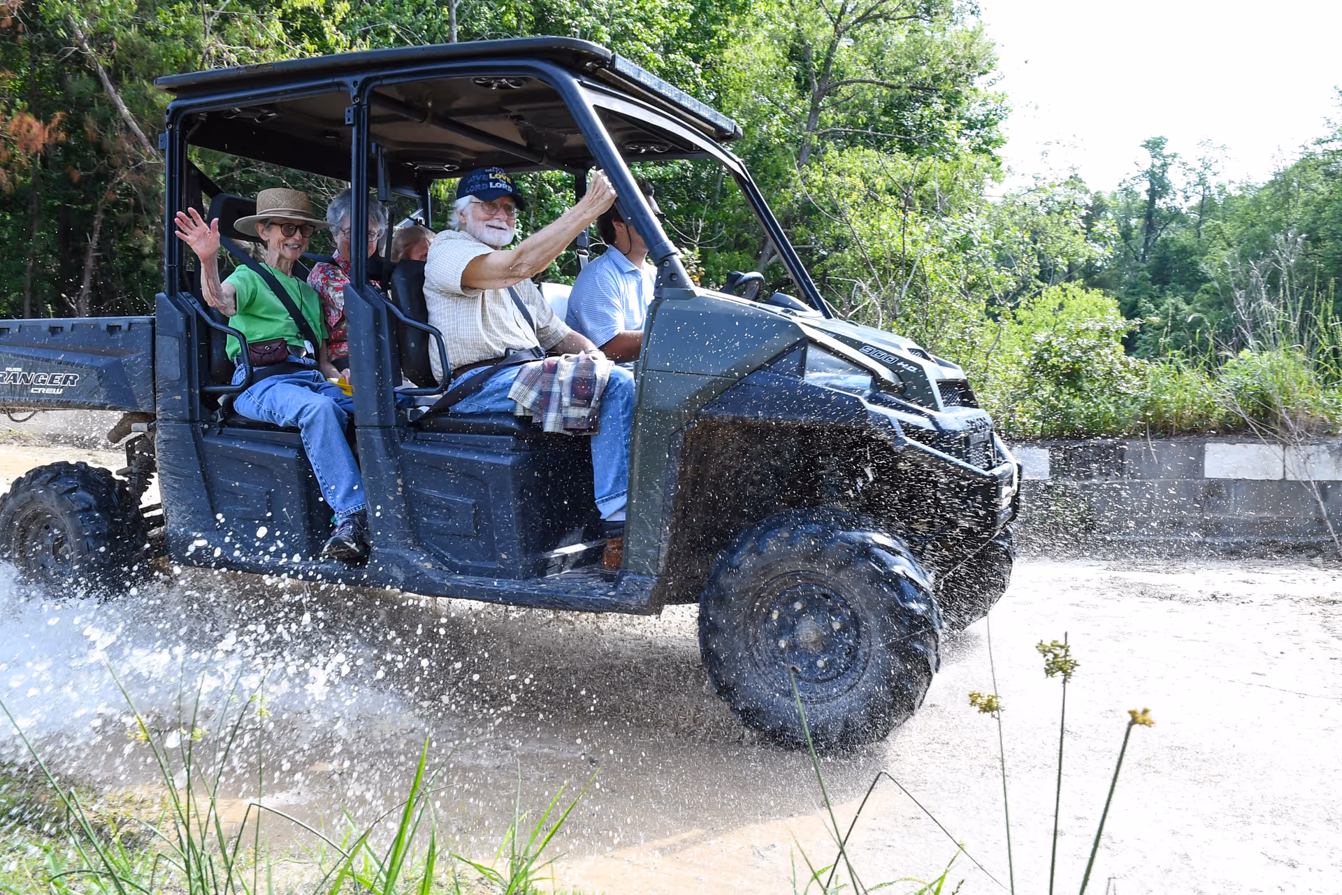 Four elderly people riding in an off-road utility vehicle splashing through a shallow puddle on a dirt path surrounded by greenery. One woman in a green shirt and hat is waving and smiling.